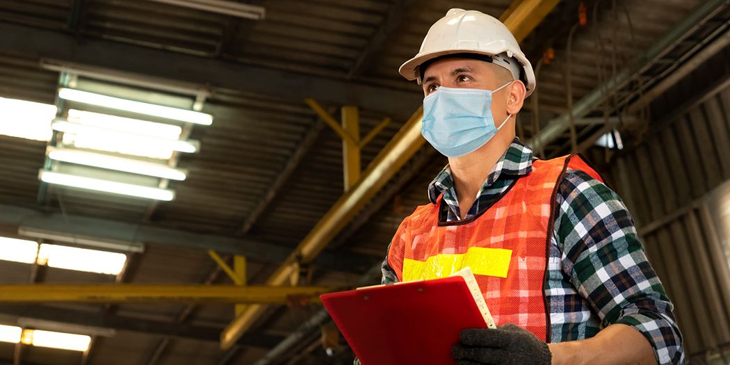 A man in a surgical mask and safety vest surveys the manufacturing floor