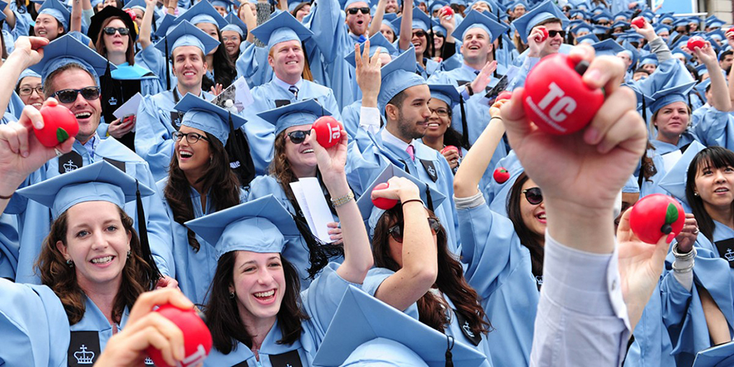 Teachers College graduates in their graduation caps and gowns hold foam apples that read 