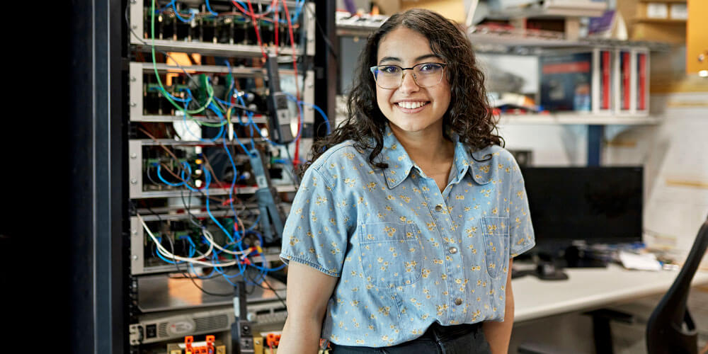 Student smiles in front of computer and machine with wires